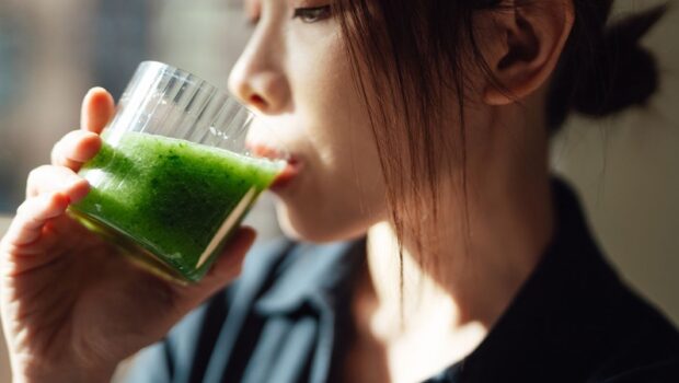 Young Asian woman drinking a glass of fresh green fruit and vegetable smoothie with natural sunlight. Healthy superfood. Detox diet.